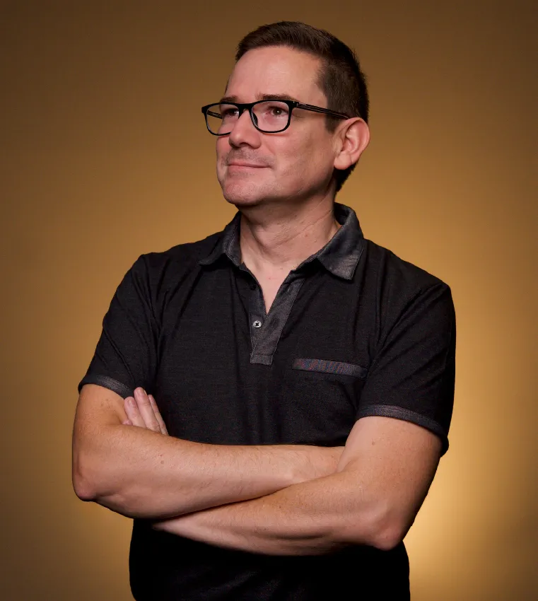Hombre con gafas y camisa negra, mirando hacia la cámara con una expresión neutral.